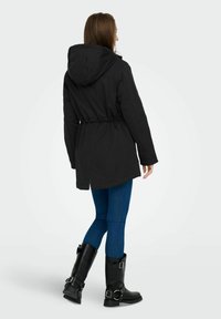 Črna parka jakna s pasom, dolgimi rokavi in kapuco. Usklajena z modrimi kavbojkami in črnimi visokimi škornji s sponkami.