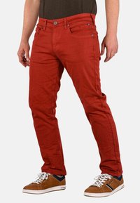 Pantalones de mezcla de algodón rojos con un corte ajustado, cinco bolsillos y cierre de botón. Combinados con zapatillas marrones con cordones y detalles en blanco.