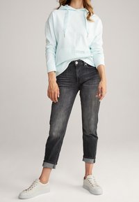 Light blue hoodie with drawstrings and front pocket, paired with dark denim, cuffed at the ankle, and white sneakers with gold accents.