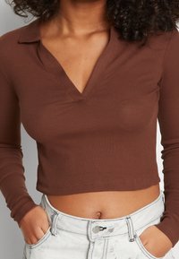Brązowy, prążkowany krótki top z długim rękawem i dekoltem w kształcie litery V oraz kołnierzykiem; dopasowany krój podkreśla talię, połączony z jasnymi, wysokimi dżinsami.