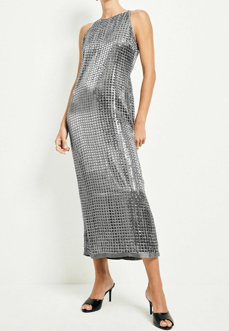 Robe midi sans manches en sequins argentés avec motif en grille, portée avec des mules à talons noires à bout ouvert sur une femme debout.