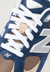 Blue suede sneaker features white laces, a beige mesh panel, and a silver logo accent. Textured materials create distinct visual contrast.