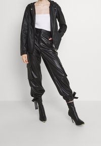 Veste en simili cuir noir avec capuche, assortie à un pantalon cargo noir doté de poches latérales et de poignets noués, ainsi qu'à des bottes à talons noires brillantes.