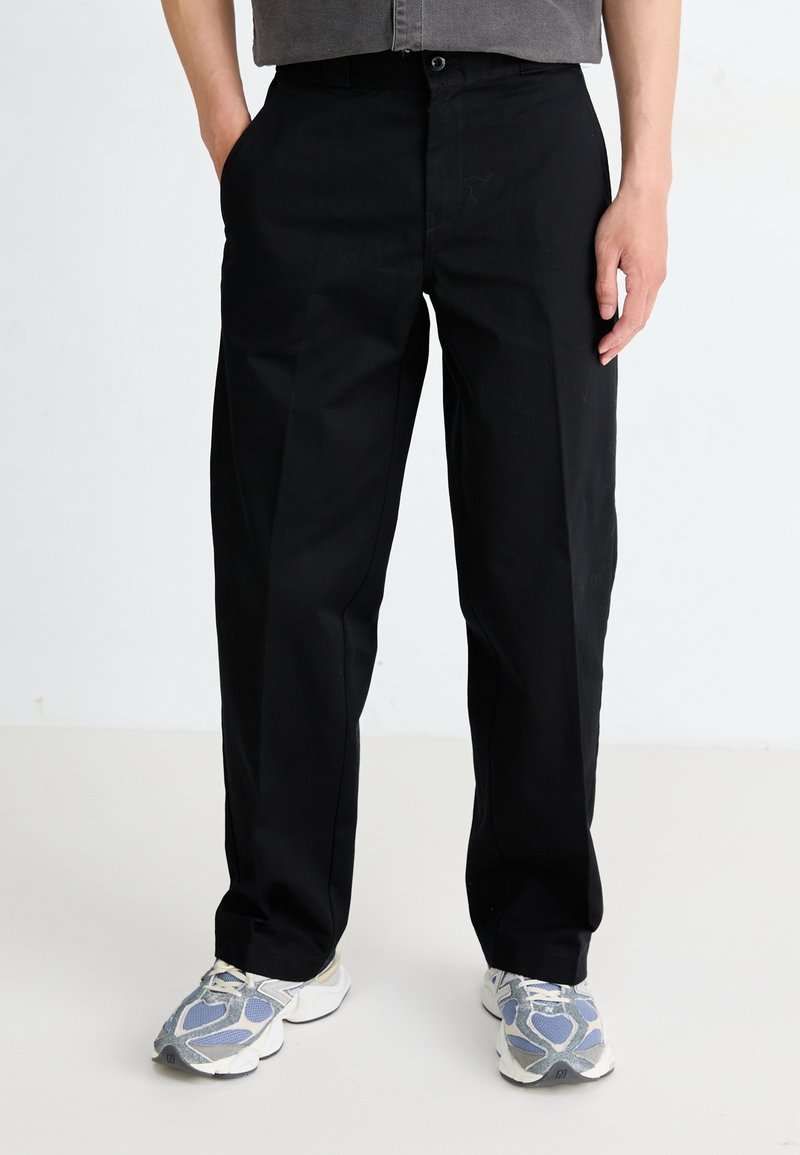 Homme portant un pantalon noir droit et des baskets grises, debout avec une main dans la poche sur un fond uni.