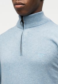 Gros plan d'une personne portant un pull en maille bleu clair avec un col demi-zip et un petit logo Barbour brodé sur la poitrine.