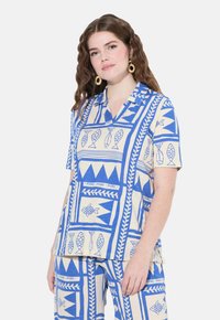 Camicia a maniche corte con colletto color crema, decorata con fantasie geometriche di pesci e foglie blu; tessuto morbido, vestibilità ampia e spacchi laterali per maggiore comfort.
