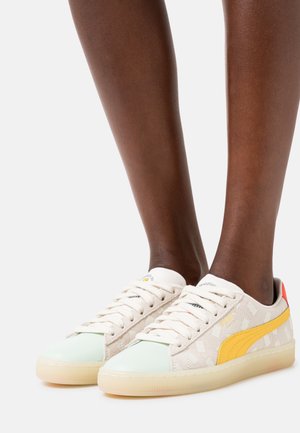 Donkere benen die beige en witte Puma sneakers dragen met een gele streep en een doorzichtige zool, tegen een witte achtergrond.