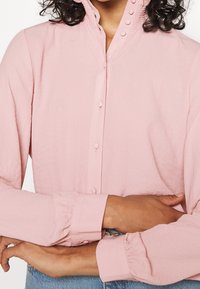Camicia rosa con colletto alto, dettagli arricciati e maniche lunghe. Presenta chiusure con bottoni e polsi arricciati. Tessuto liscio.