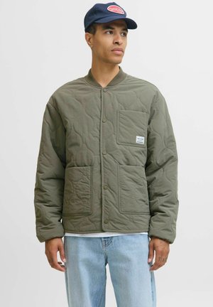 Quilted olive green jacket with snap closures, two front pockets, and a small branded patch; worn over a white shirt and light jeans.