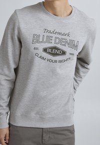 Sudadera gris con cuello redondo, puños y dobladillo acanalados, y un diseño de logo impreso en negro, con detalles de texto en la parte delantera.