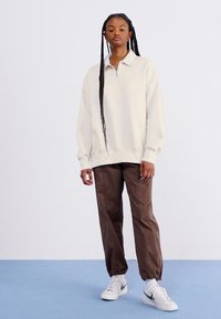 Κρέμ μισό-φουτερ πουκάμισο σε συνδυασμό με καφέ κομμένα παντελόνια και λευκά αθλητικά παπούτσια, με χαλαρή εφαρμογή και μινιμαλιστικό σχέδιο.