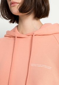 Jasnoróżowa bluza z kapturem wykonana z miękkiego materiału, z kieszenią typu kangur i sznurkiem do regulacji. Klasyczny fason z ribbowymi mankietami.