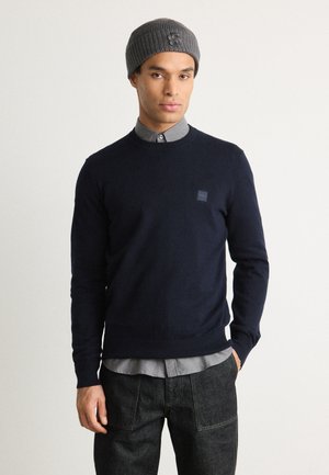 Navy knitted sweater, crew neckline, small logo patch. Worn over a light grey collared shirt and paired with dark denim pants.