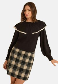 Pulover negru cu un tiv alb volanat la decolteu și mâneci lungi bufante, asortat cu o fustă în carouri în tonuri de bej și negru.