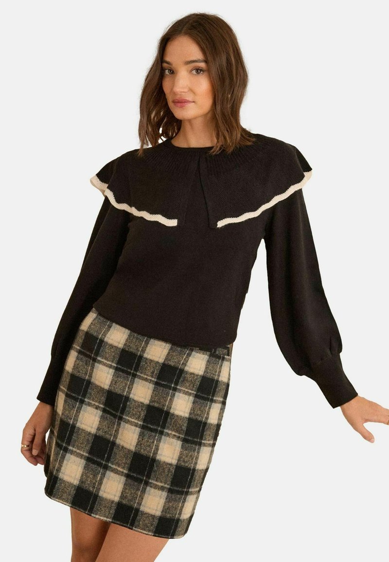 Pulover negru cu un tiv alb volanat la decolteu și mâneci lungi bufante, asortat cu o fustă în carouri în tonuri de bej și negru.