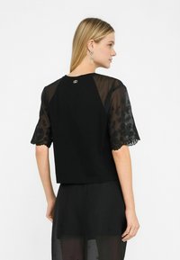 Top nero con maniche in pizzo trasparente a motivo floreale e orlo scallop. Design semplice con un piccolo emblema sullo scollo posteriore.