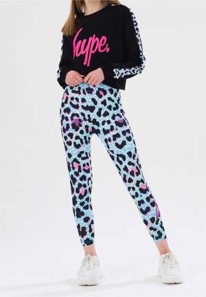 Kvinde iført sort cropped sweatshirt med lyserød "hype" tekst og matchende leopardprint leggings i blå, pink og sort, med hvide sneakers.