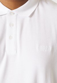 Zbliżenie na białą teksturowaną koszulkę polo z trzema guzikami i subtelnym wytłoczonym logo "JOOP! Jeans" na piersi.