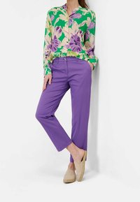 Chemise à boutons à motif floral en vert, violet et crème ; associée à un pantalon violet fuselé. Le mannequin porte des chaussures beige à enfiler. Tissu lisse et léger.