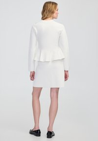 Chaqueta blanca de peplum con mangas largas y un diseño ajustado, combinada con una falda blanca a juego. La modelo lleva zapatos planos negros.