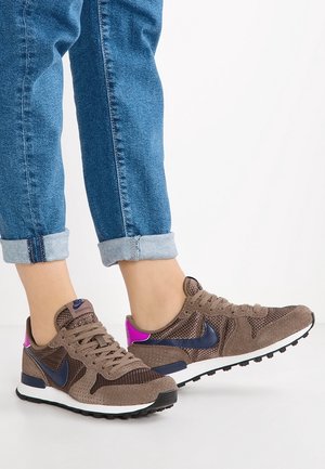 Baskets Nike marron avec des accents bleu marine et rose portées avec un jean bleu retroussé sur fond blanc.
