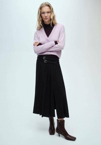 Ljuslila kavaj över en mörk polotröja, i kombination med en svart plisserad kjol med dubbla spännen, och bruna ankelstövlar.