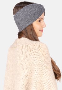 Un bandeau en laine grise avec un design torsadé, porté par une personne aux cheveux longs et bruns, associé à un cardigan tricoté de couleur beige clair.