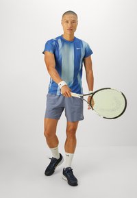 Joueur de tennis masculin en chemise à rayures bleues et en shorts gris tient une raquette de tennis, se préparant à frapper contre un fond uni.