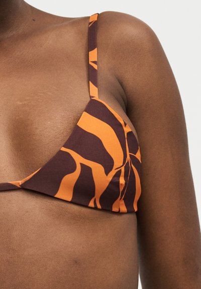 Κοντινό πλάνο ενός ατόμου που φοράει μπικίνι πάνω μέρος με καφέ και πορτοκαλί σχέδιο και λεπτές τιράντες σε μαυρισμένο δέρμα με απλό φόντο.