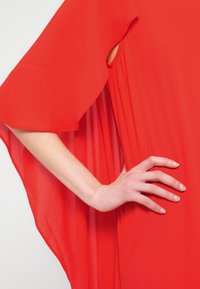 Κόκκινο μεταξωτό μπλουζάκι με φαρδιά, ροή μανίκια, πλαϊνή σχισμή και πιετέ λεπτομέρειες. Το ύφασμα έχει ελαφριά, διαφανή υφή.