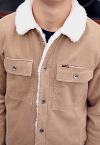 Beżowa kurtka z korduroyu z białym kołnierzem sherpa, dwoma kieszeniami na piersi z zapięciem na napy oraz metką na lewej kieszeni.
