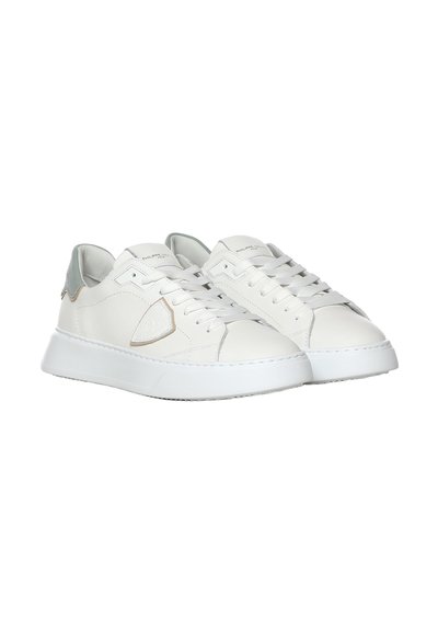 Paire de baskets basses blanches avec des accents gris, lacets à l'avant, semelles épaisses et un emblème en forme de bouclier sur les côtés.