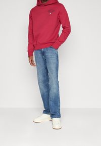 Czerwona bluza z kapturem z logo, zestawiona z jasnoniebieskimi, prostymi dżinsami i białymi niskimi tenisówkami, na jednolitym tle.