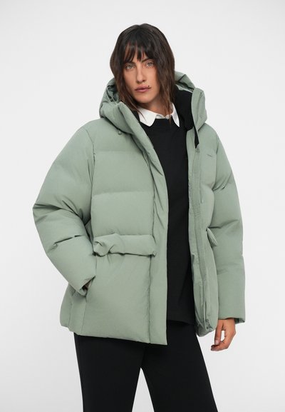 Padded green puffer jacket with a hood, featuring a zip closure, front pockets, and a smooth, matte texture. Worn over a black top.