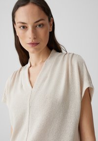 Maglione di un beige chiaro con scollo a V, maniche corte, tessuto testurizzato e dettagli con cuciture sottili, progettato per una vestibilità morbida.
