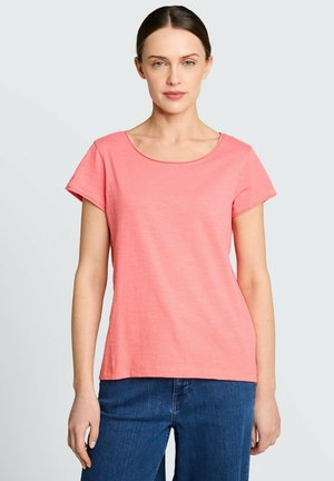 Femme aux cheveux foncés portant une chemise corail à manches courtes et un jean bleu, debout devant un fond uni gris clair.