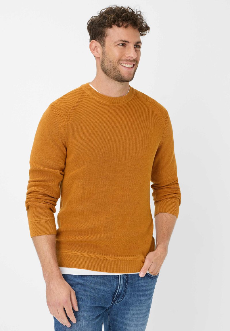 Senfgelbes Strickpullover mit gerippter Textur, rundem Ausschnitt und langen Ärmeln, kombiniert mit Jeans.