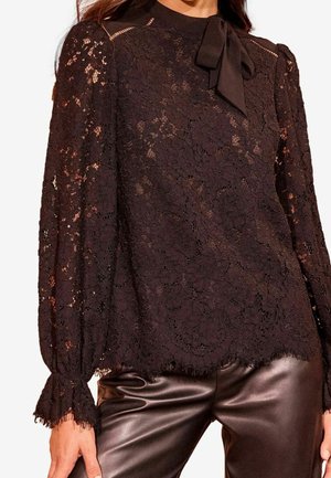 Zwarte kanten blouse met lange mouwen, bloemenpatroon en neklint, gecombineerd met donkerbruine leren broek.