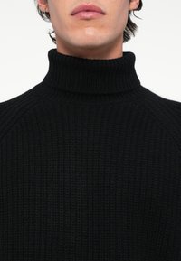 Homme portant un pull à col roulé noir côtelé, montrant le cou et le bas du visage sur un fond blanc uni.