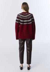 Pull en tricot rouge avec un design fair isle noir, blanc et vert ; associé à un pantalon texturé marron et des chaussures noires.