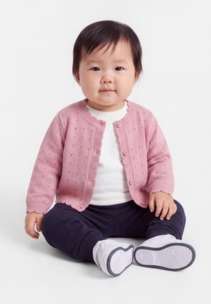 Tout-petit assis, portant un cardigan rose tricoté, une chemise blanche, un pantalon sombre et des chaussures blanches, sur un fond clair uni.