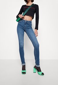 Niebieskie wąskie dżinsy z delikatnym wyblaknięciem, połączone z czarnym, krótkim topem z długim rękawem oraz zielonymi sandałami na słupku z szerokimi paskami.