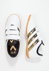 Zapatillas deportivas blancas con tres acentos de rayas negras y doradas, correas de Velcro, parte superior texturizada y suela de goma.