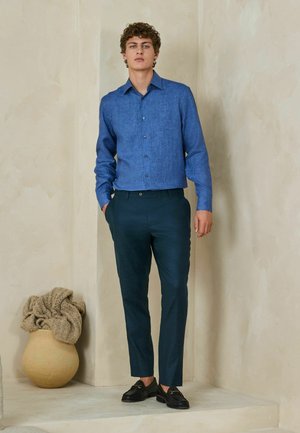 Young man standing indoors wearing a blue button-up shirt, dark pants, and black loafers, near a beige textured wall and pottery with fabric.