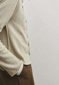 Cardigan beige con bottoni, maniche lunghe, tessuto testurizzato e orlo sportivo. Indossato sopra una camicia bianca con polsino visibile.
