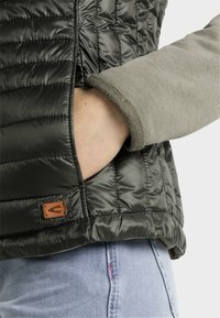 Mörkgrön vadderad jacka med en slät textur, som har en sidoficka och en logotyp i brunt läder. Ärmarna är av grått tyg.