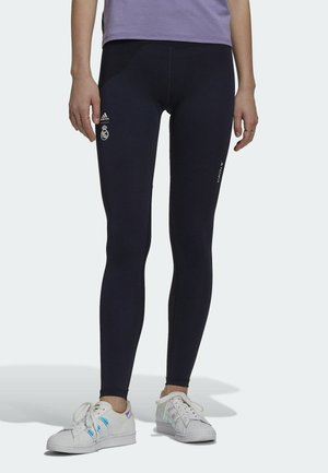 Osoba nosząca czarne legginsy Adidas z logo Realu Madryt i białe sneakersy, stojąca na jednolitym tle.