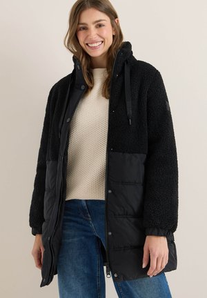 Doudoune noire avec une partie supérieure en polaire texturée, fermetures éclair et pression, capuche ajustable et poches latérales. Portée sur un pull crème.