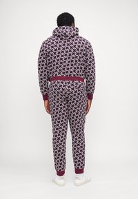 Hooded trainingspak in donkerpaars met een geometrisch patroon in crème en bordeaux. Voorzien van ribgebreide manchetten en tailleband, gecombineerd met witte sneakers.