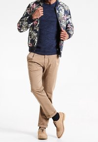 Floral bomber jacket with navy background, textured knit sweater, beige trousers, and light brown shoes with brogue detailing.